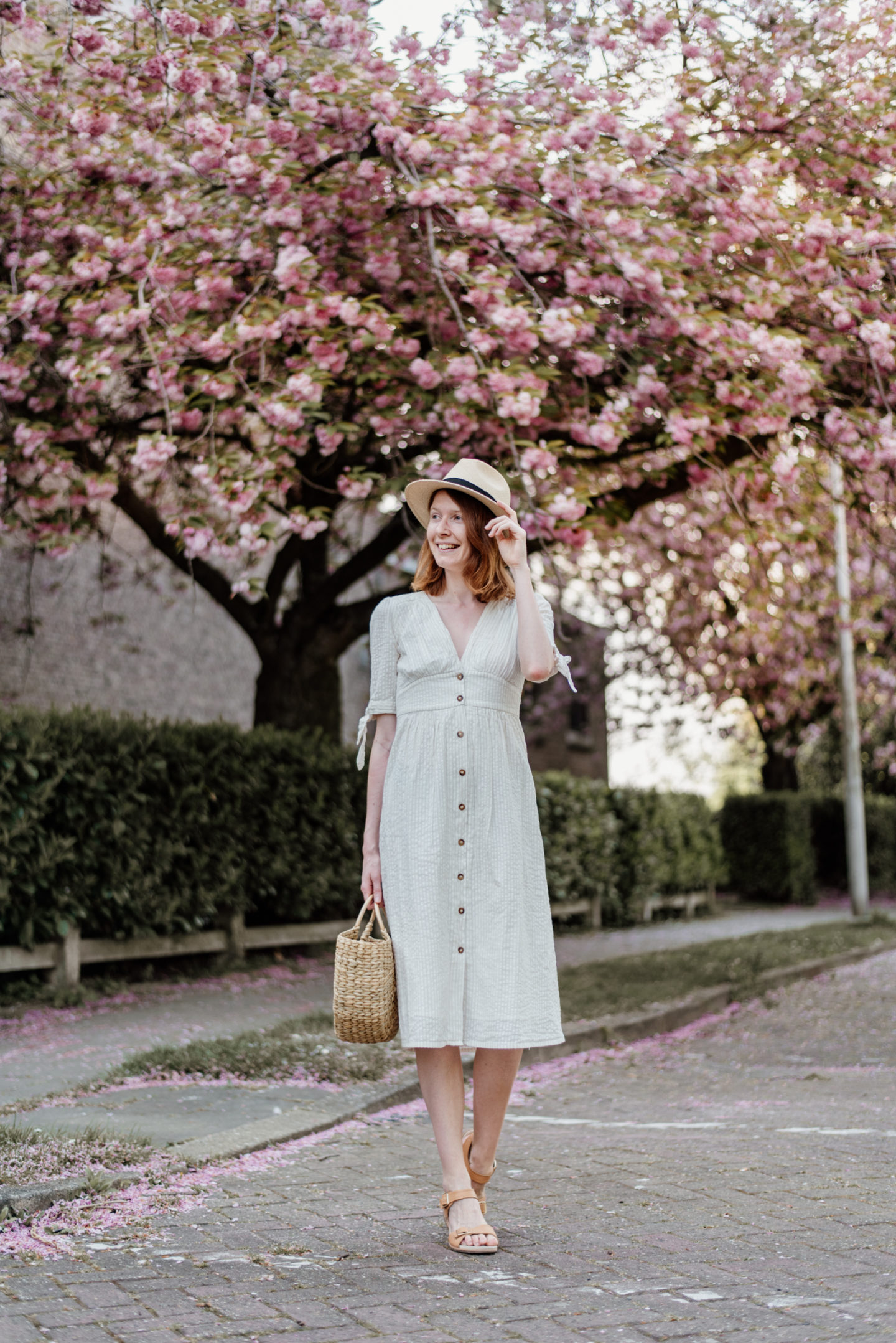Girl in white dress and with a straw bag posing in front of cherry blossoms