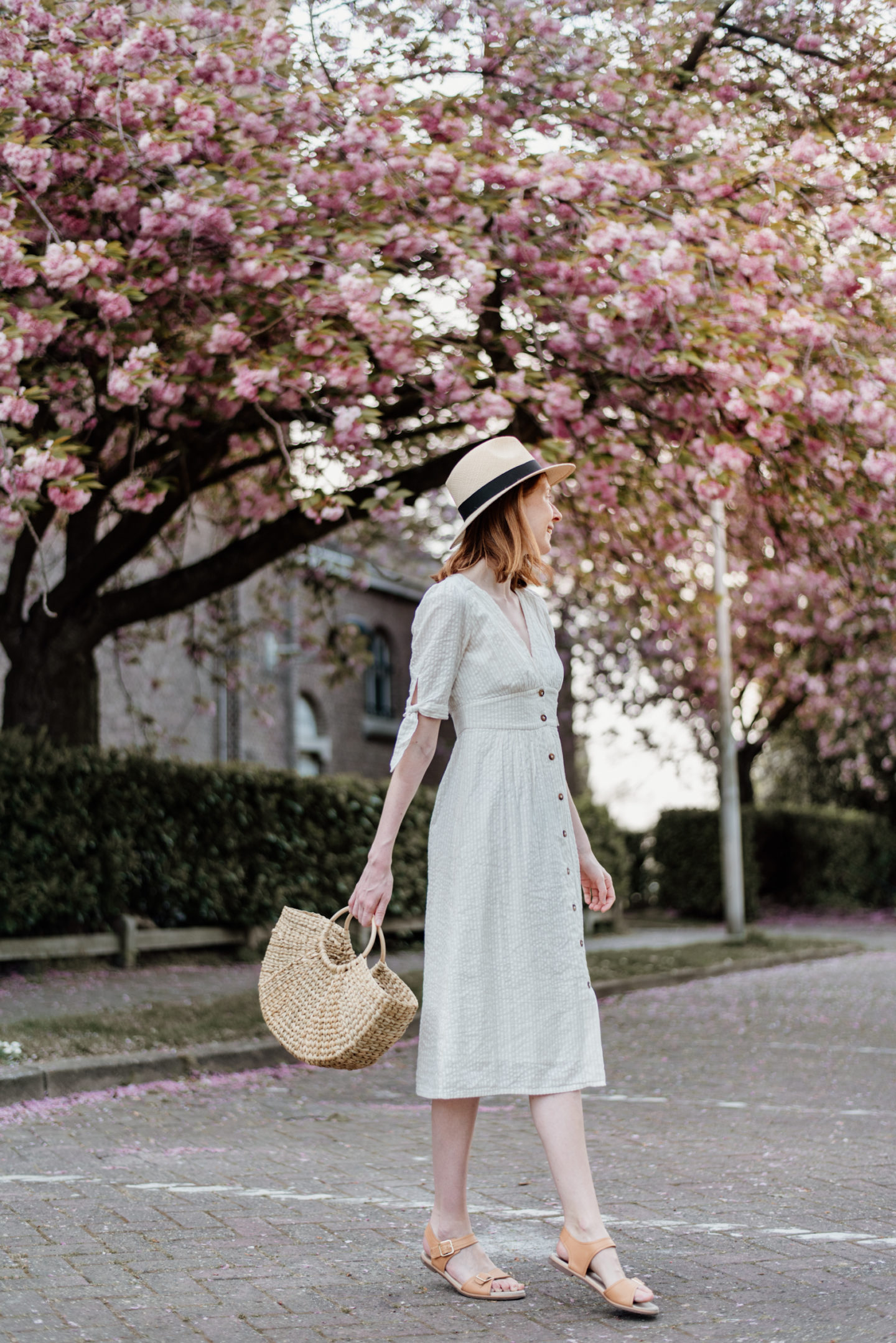 Girl in white dress and with a straw bag posing in front of cherry blossoms