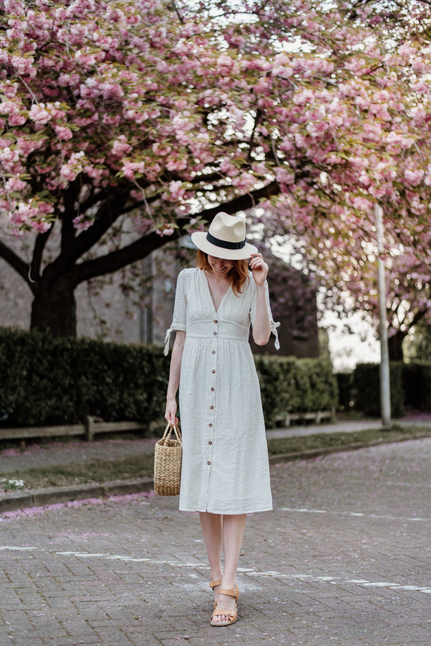 Girl in white dress and with a straw bag posing in front of cherry blossoms