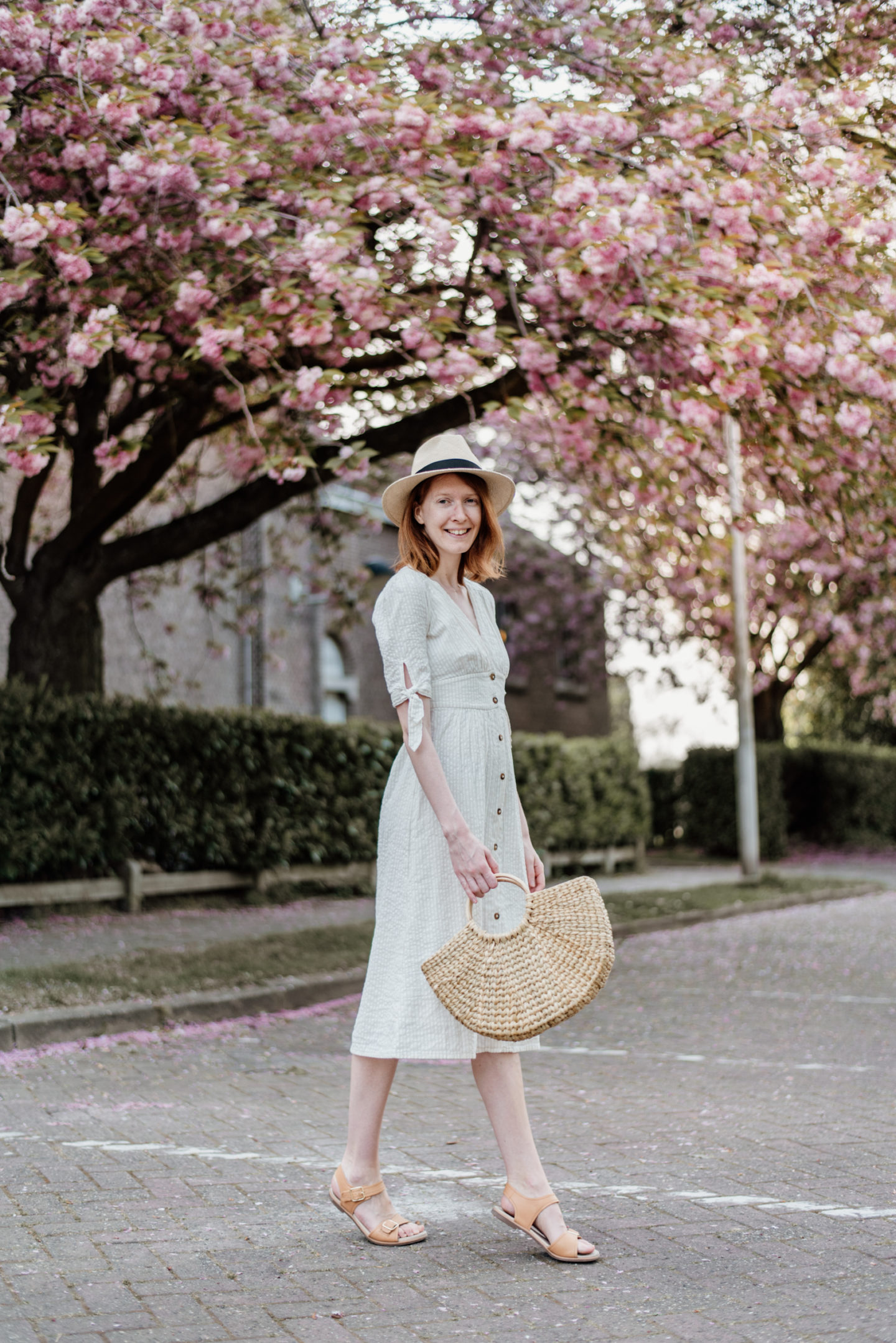 Girl in white dress and with a straw bag posing in front of cherry blossoms